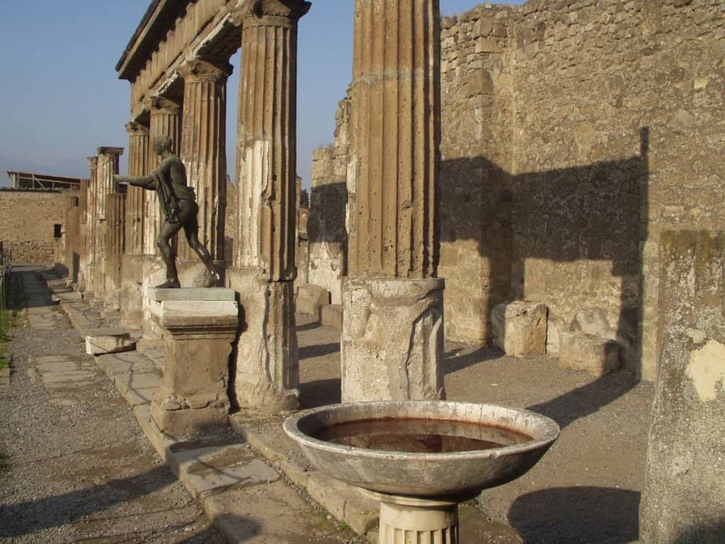 Ancient Roman ruins with stone columns, a statue, and a stone basin in an outdoor setting.