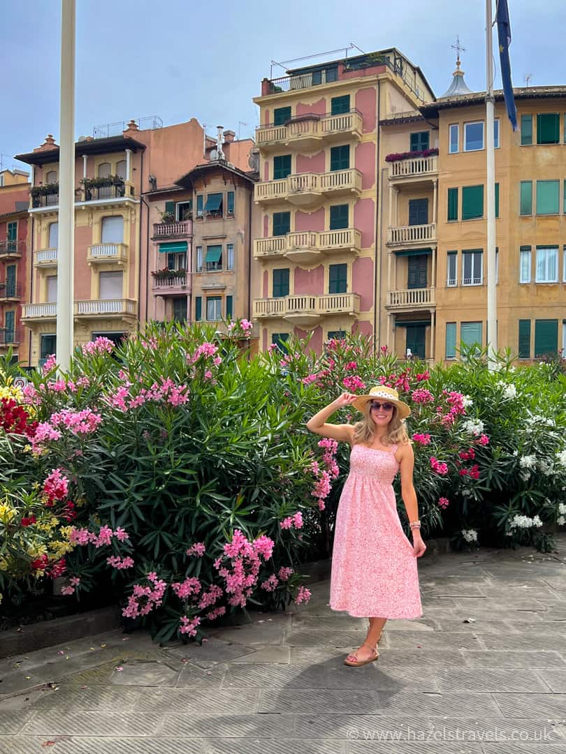 Woman in a pink dress and sun hat posing beside vibrant flowering bushes with colourful buildings in Santa Margherita Ligure, Italy.