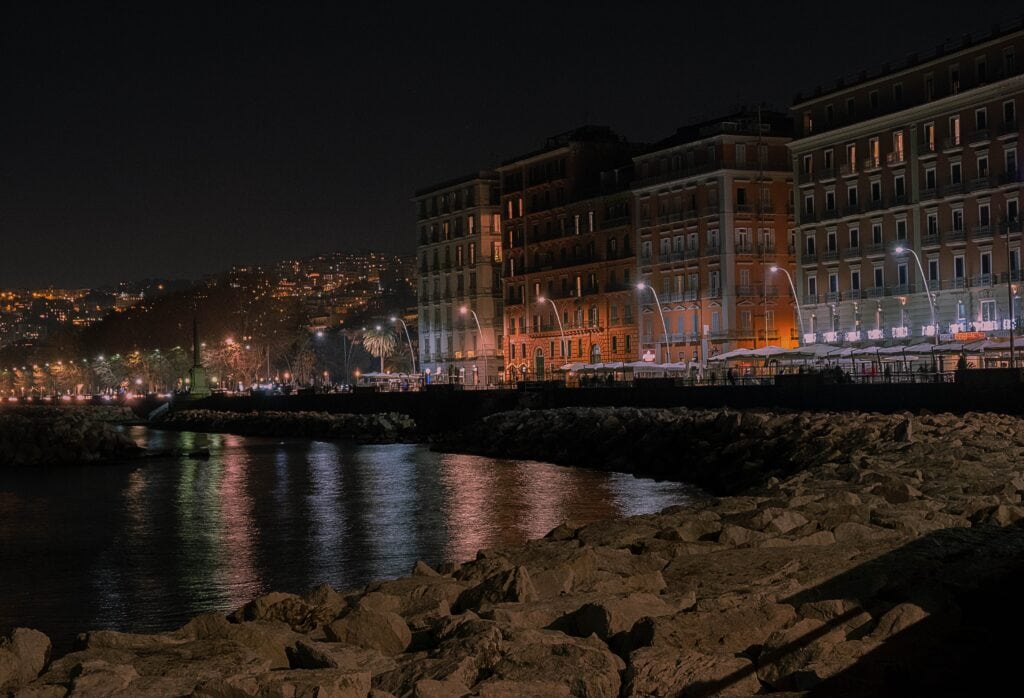 Night view of a waterfront cityscape with illuminated buildings lining the shore, reflections in the water, and rocky foreground.