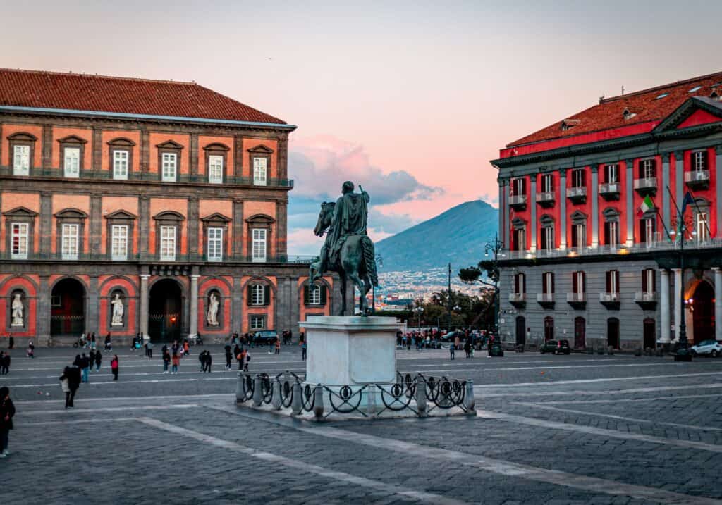A statue of a rider on horseback stands in a large plaza surrounded by historic buildings, with a mountain in the background under a pink and blue sky.