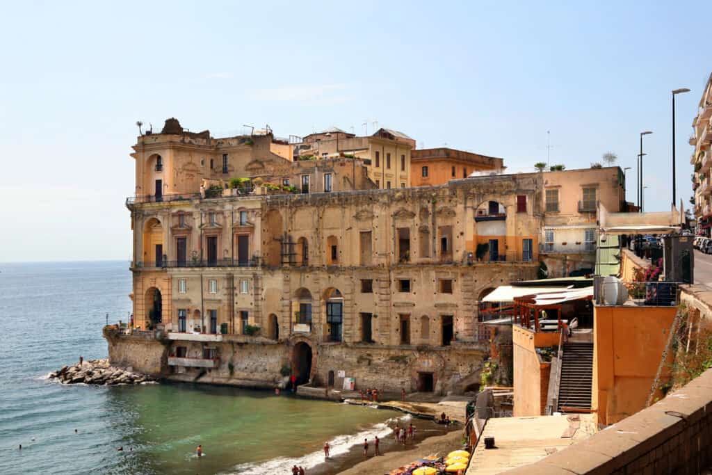 Historic waterfront building with weathered facade by a small beach, overlooking a calm sea. People are swimming and sunbathing. Urban setting with adjacent street on the right.