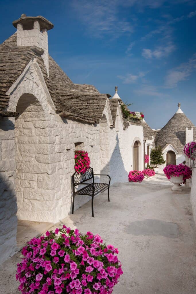 White stone buildings with conical roofs line the charming streets of Alberobello, featuring pink flowers in pots along a pathway with a black metal bench, all under a clear blue sky.