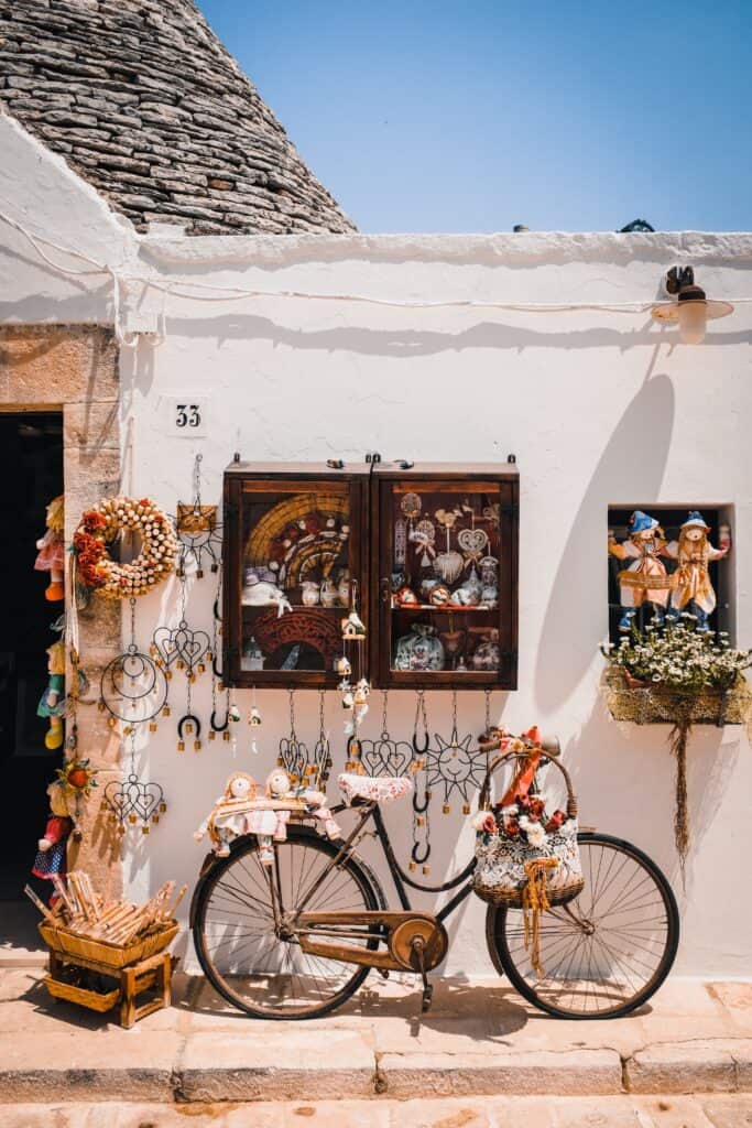 A vintage bicycle with a basket full of decorative items is parked against a white building in charming Alberobello. The wall is adorned with various hanging crafts and ornaments.