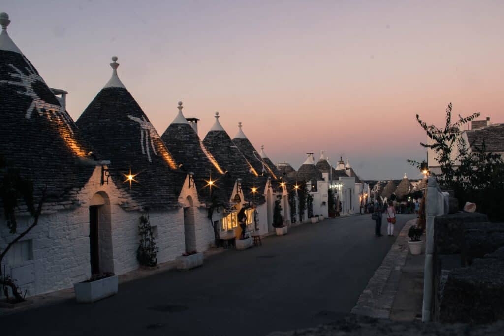 A street in Alberobello lined with traditional conical-roofed buildings under a dusk sky. People stroll along the sidewalk, and warm lights softly illuminate the charming structures.