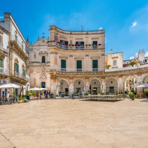 A picturesque town square in Martina Franca features historic white buildings, arched walkways, and outdoor seating under umbrellas, set against the clear blue sky of Puglia.