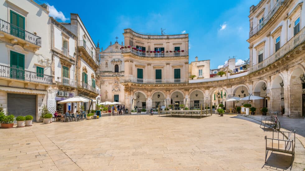 A picturesque town square in Martina Franca features historic white buildings, arched walkways, and outdoor seating under umbrellas, set against the clear blue sky of Puglia.