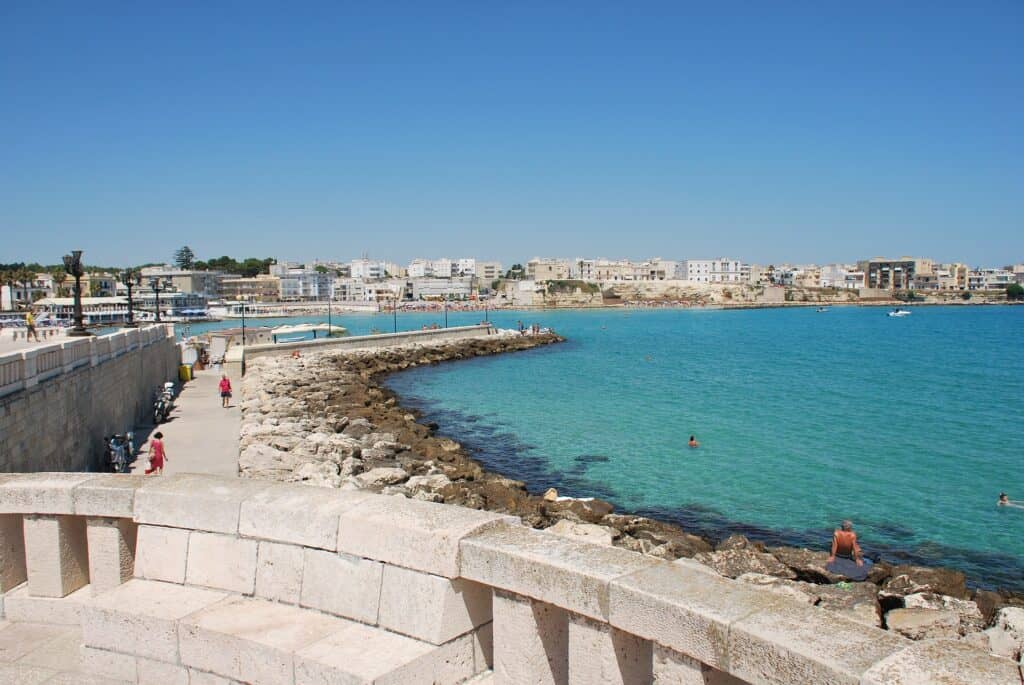 Coastal town view in Otranto with a curved stone pier, clear blue water, and several people enjoying a sunny day along the shoreline. Houses and buildings are visible in the background.