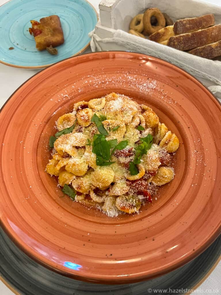 At Alberobello restaurant, a plate of pasta crowned with grated cheese and herbs sits elegantly on a red plate. In the background, a blue plate cradles a piece of bread alongside a basket filled with an assortment of breads.