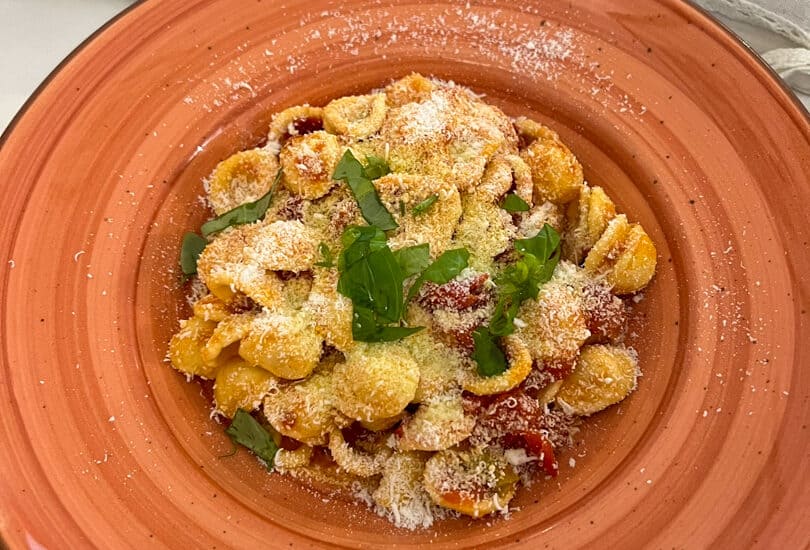 At Alberobello restaurant, a plate of pasta crowned with grated cheese and herbs sits elegantly on a red plate. In the background, a blue plate cradles a piece of bread alongside a basket filled with an assortment of breads.