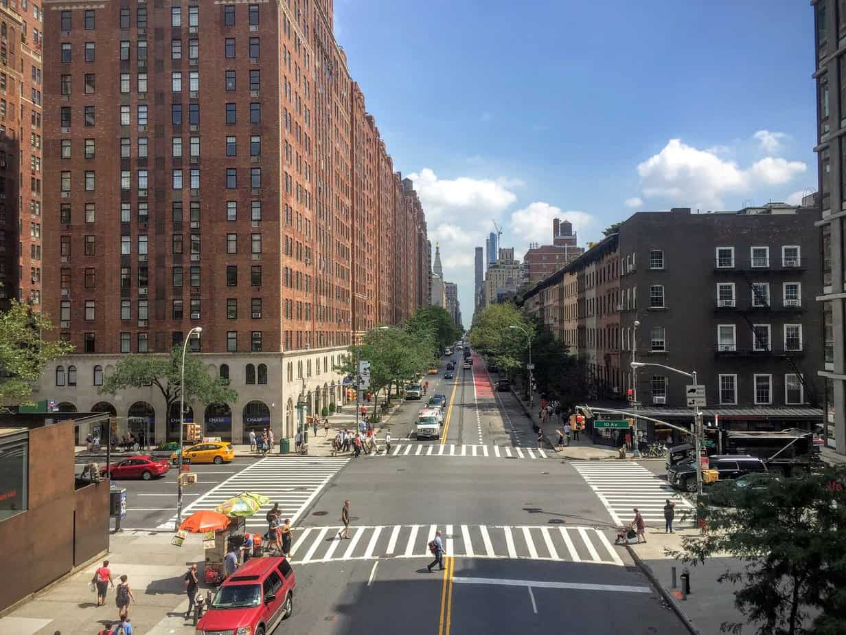 A bustling city street intersection with tall brick buildings on both sides, as seen from the Highline Park NYC, showcases pedestrians crossing and cars moving below under a clear blue sky.