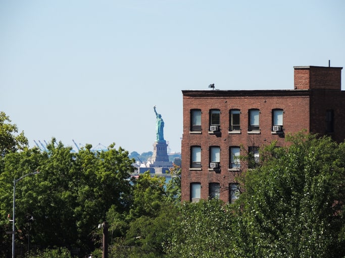 From the vantage point of the High Line, the view features the Statue of Liberty in the distance, partially obscured by trees and a brick building in the foreground.