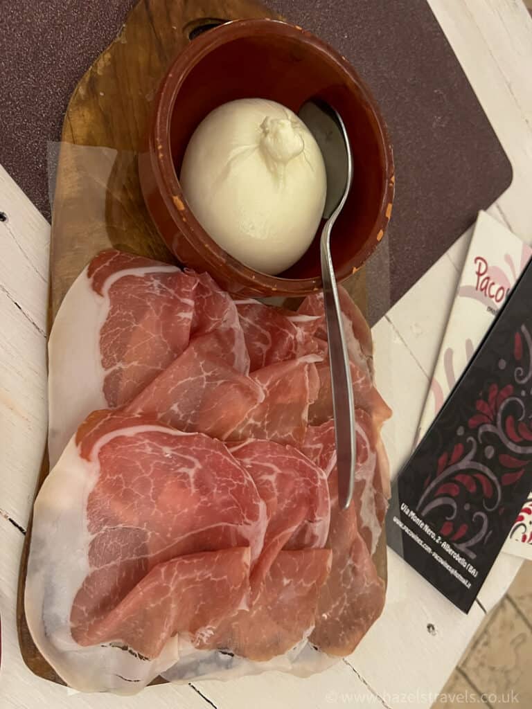 Sliced prosciutto and a ball of mozzarella rest in a wooden bowl on a cutting board, accompanied by a spoon, evoking the rustic charm of an Alberobello wine bar.