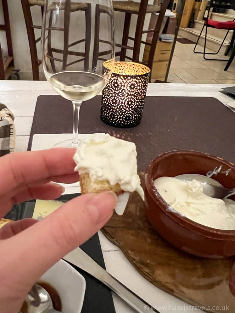 A hand holds a slice of bread with creamy cheese spread beside a bowl of cheese, a glass of white wine, and a lit candle on an intimate Alberobello wine bar table.