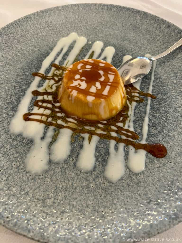 A caramel flan dessert on a blue plate, garnished with white and brown sauce, with a spoon placed beside it.