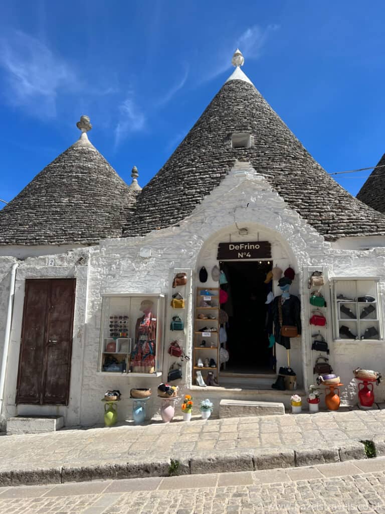 In the charming alley, a stone building with conical roofs reminiscent of Alberobello proudly displays an array of hats and bags outside.