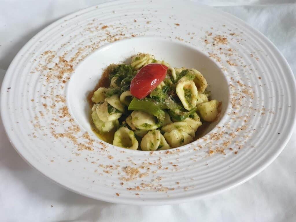 A white plate of orecchiette pasta with broccoli rabe, topped with a cherry tomato and a sprinkling of breadcrumbs.