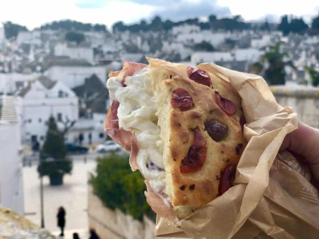 A hand holds a focaccia sandwich filled with creamy white cheese and prosciutto, wrapped in paper, against a background of a white, townscape with trees.