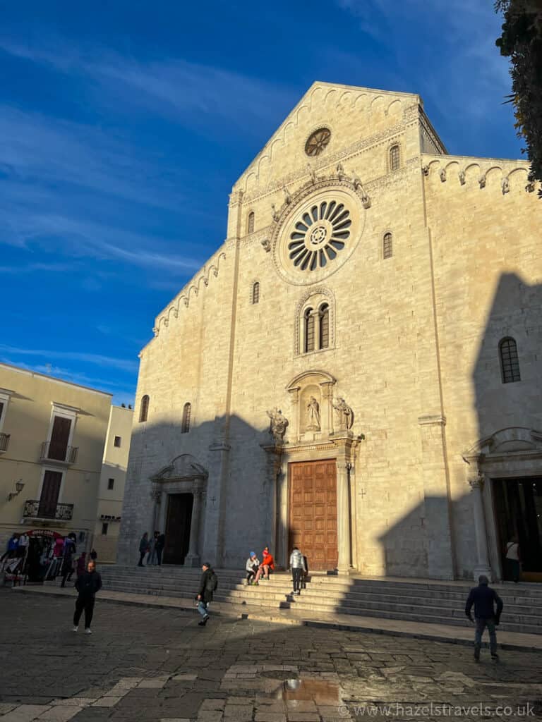 People walk in front of a historic stone church with a large rose window and a wooden door under a blue sky.