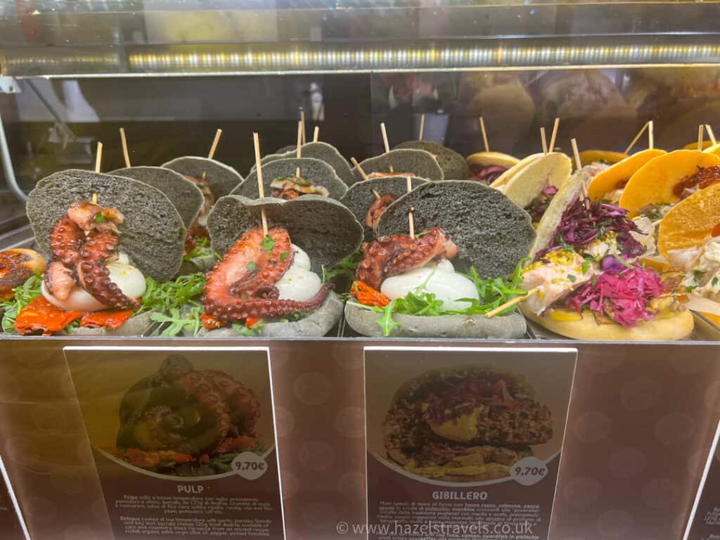 Display case with black buns filled with octopus tentacles and white toppings, garnished with greens and tomatoes. Price tags visible below.