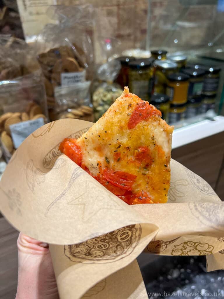 A slice of tomato-topped pizza is held in a hand, wrapped in patterned paper. In the background, shelves display jars and bags of food items.