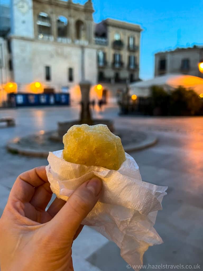 A hand holds a pastry wrapped in a napkin with a historic plaza in the background during dusk.