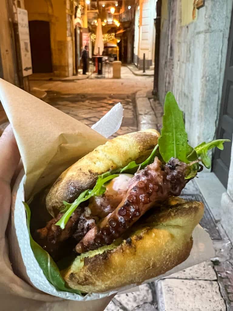 Hand holding a sandwich with grilled octopus and arugula in a pita, on a narrow, dimly lit street with outdoor seating and lights in the background.