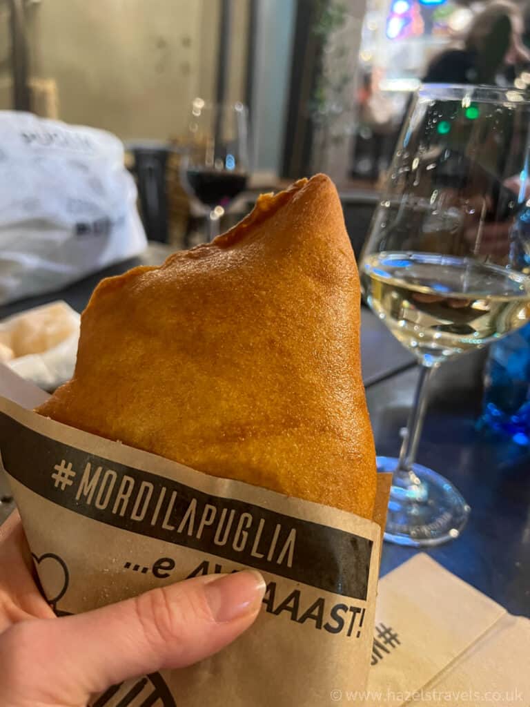 Hand holding a wrapped fried snack, with glasses of red and white wine on a table in the background.