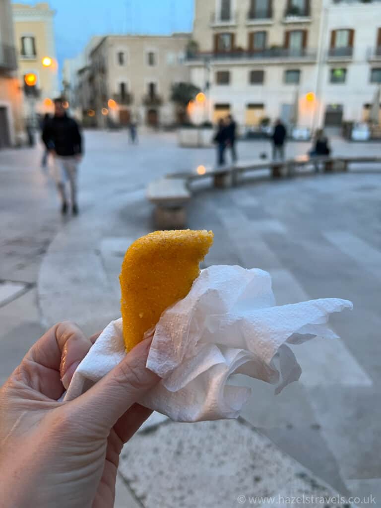 Hand holding a fried pastry wrapped in a napkin, with a blurred urban square and a few people walking in the background.