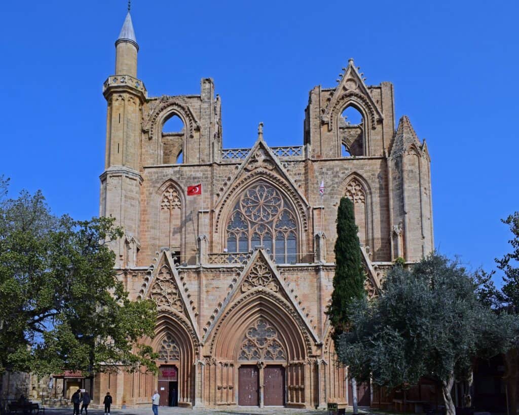 A Gothic-style stone cathedral with pointed arches and large stained glass windows, featuring a minaret and Turkish flags, stands near the Famagusta border crossing, set against a clear blue sky.