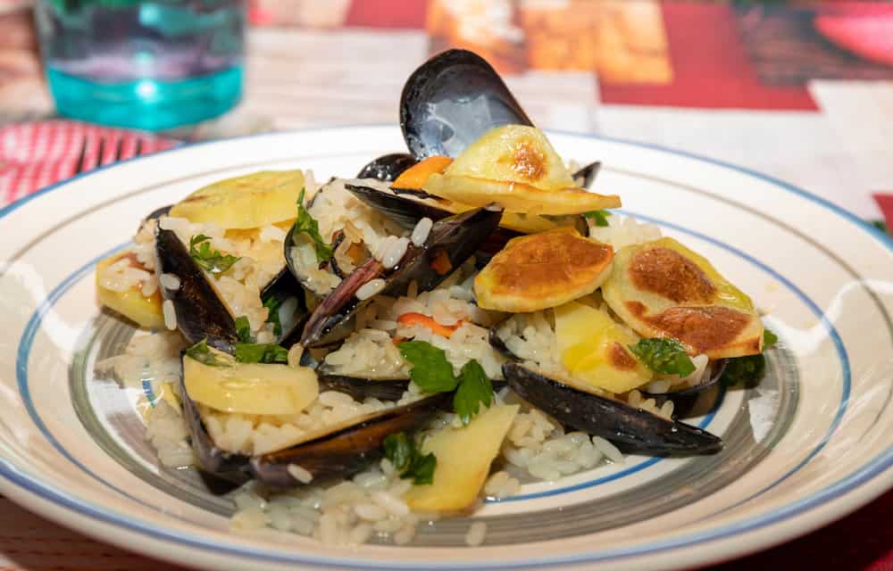 A plate of Tiella di Riso with mussels, sliced potatoes, and herbs, served on a white dish with a blue rim.