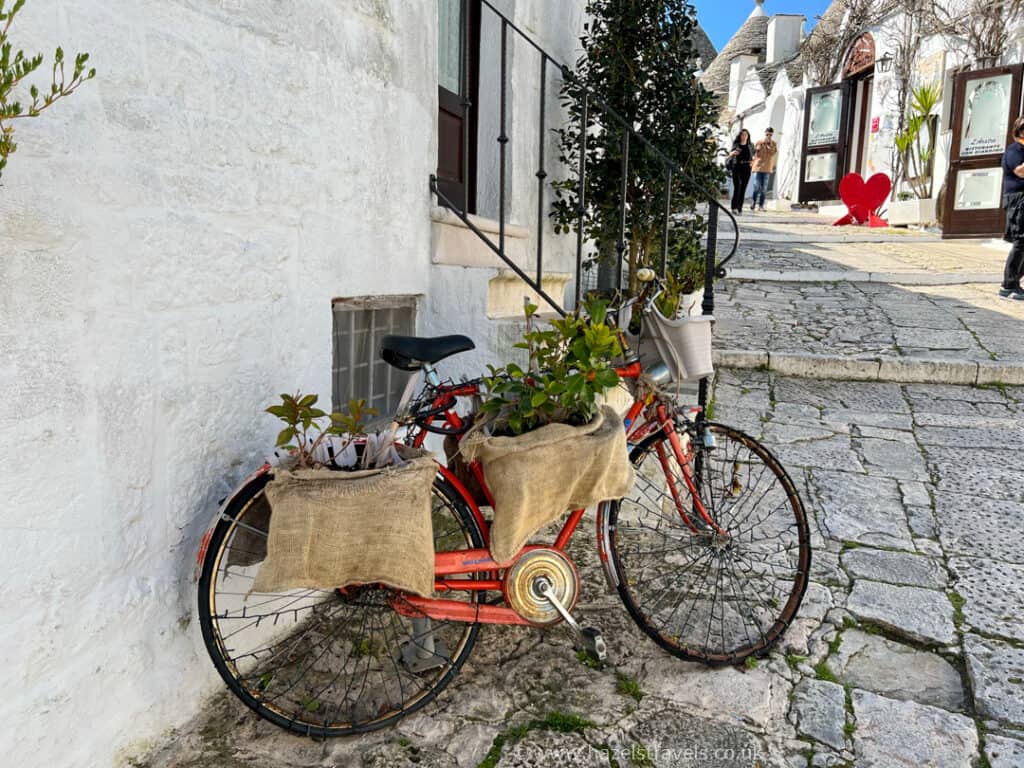 A red bicycle leaning against a white wall on a stone-paved street, with plants in burlap bags on the front and back.