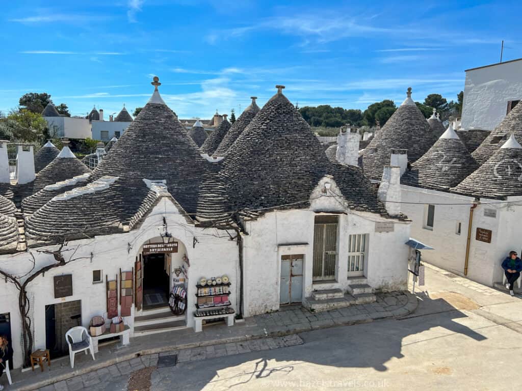 White stone houses with conical roofs under a clear blue sky. A few items are displayed outside a shop. A person stands on the right. Trees are visible in the background.