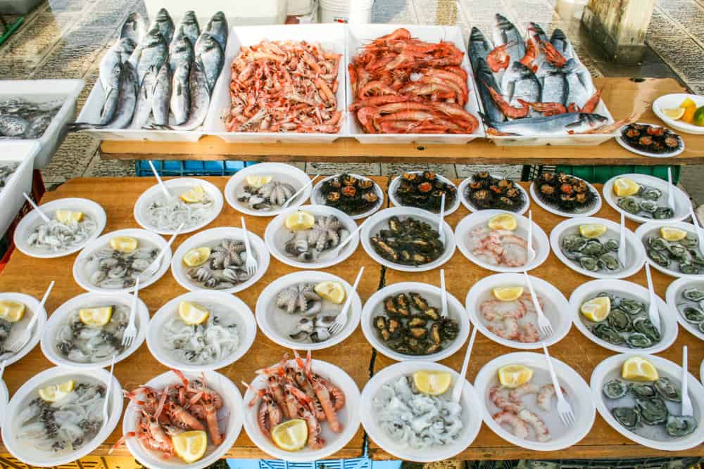 Plates of assorted seafood, including shrimp, octopus, and shellfish, displayed on a market table with lemon wedges.