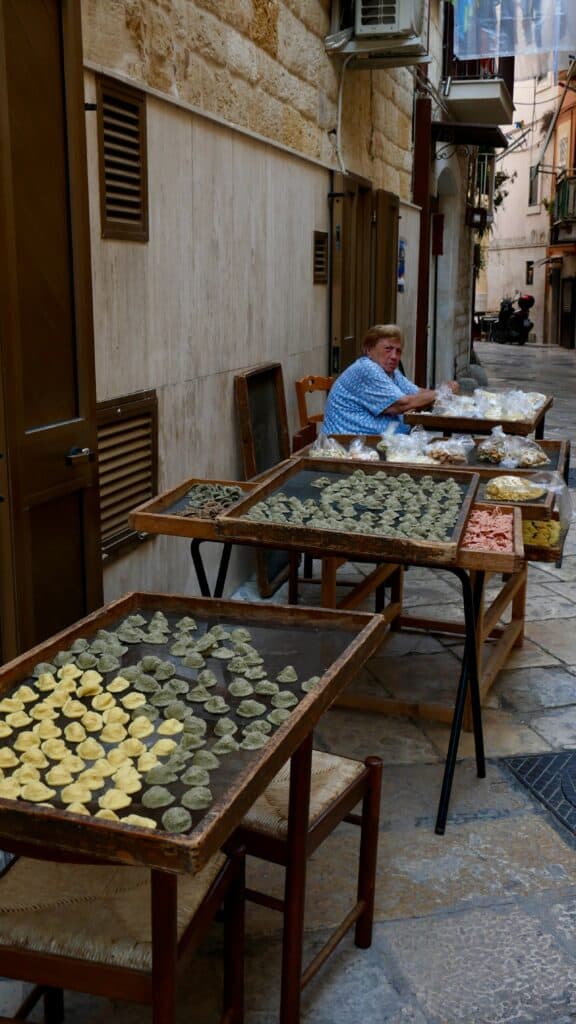 Person sitting at a table in a narrow cobblestone alley, displaying various types of dumplings on wooden trays. Stone walls and an air conditioning unit are visible in the background.