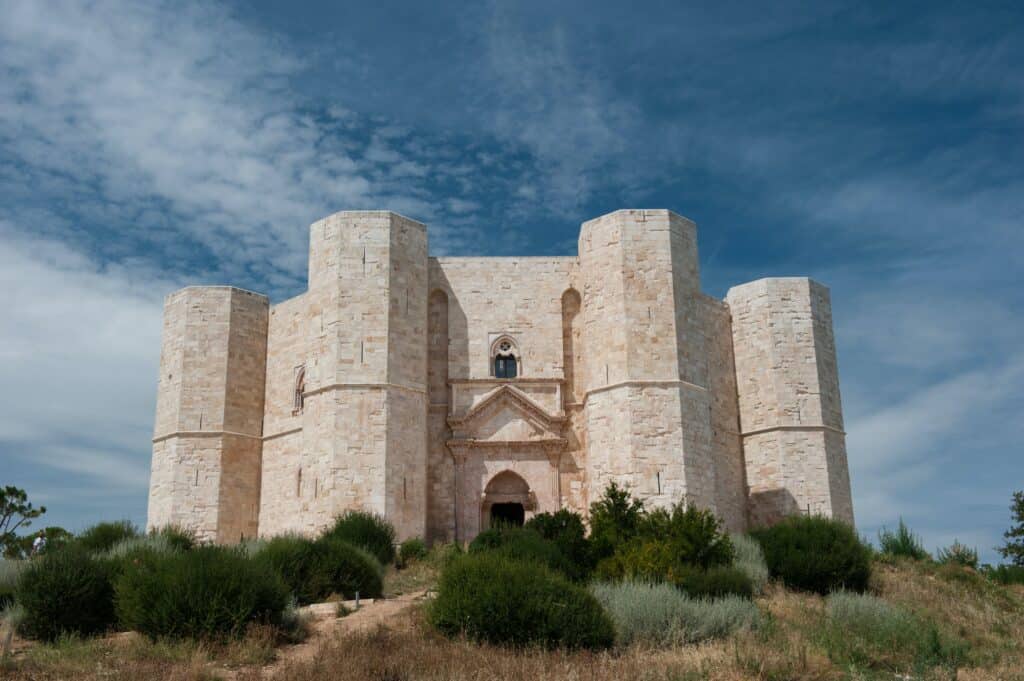 Castel del Monte, a medieval stone fortress with an octagonal shape, stands under a partly cloudy sky surrounded by the lush greenery of Puglia, echoing the charm of Alberobello's unique landscape.