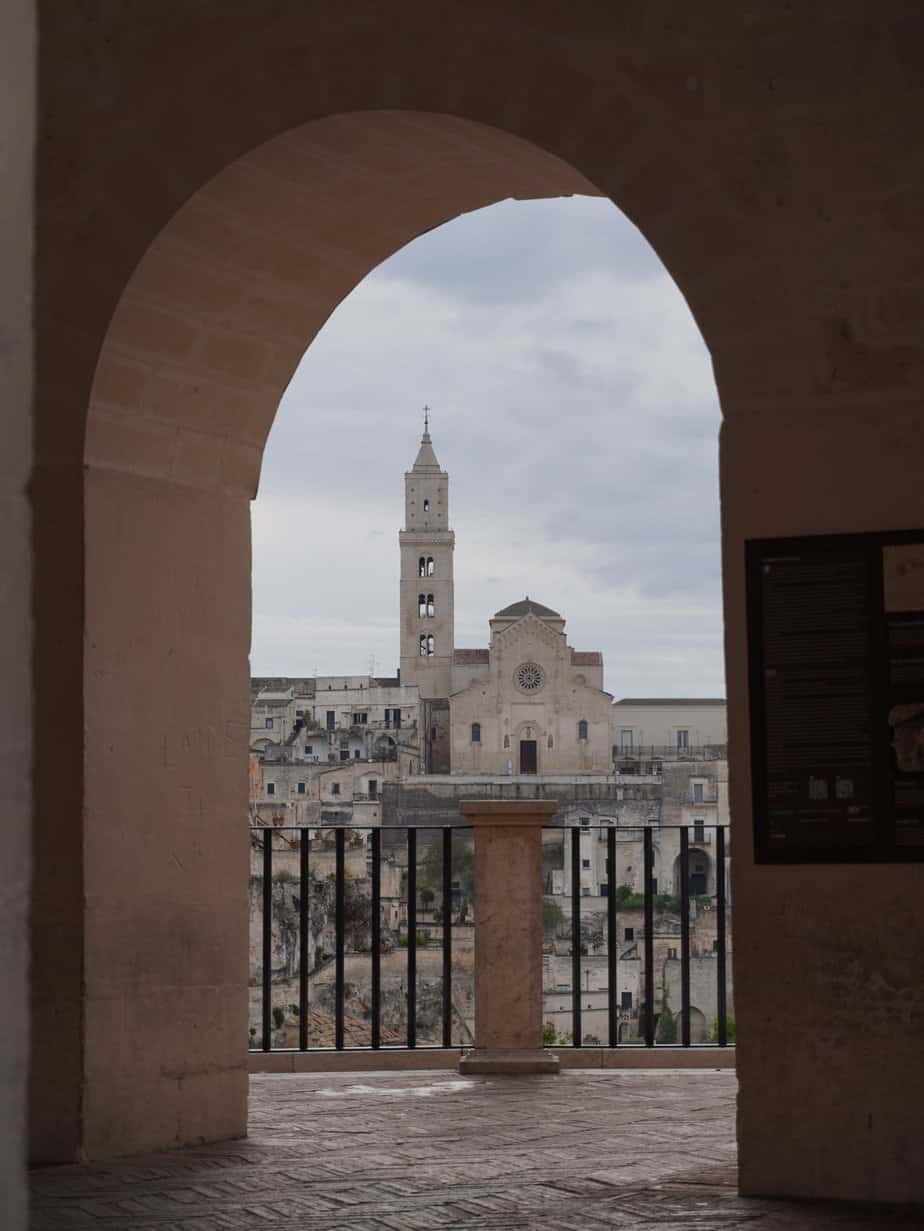 View through an archway of a historic stone building with a tall bell tower, set against a cloudy sky. A railing runs parallel to the arch.