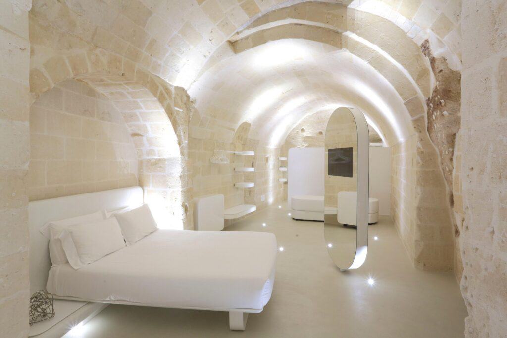 Modern bedroom in a stone cave with white furniture, including a bed and shelves. Curved stone walls and ceiling with soft lighting create a minimalist and serene atmosphere.