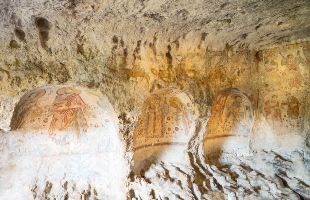 Ancient cave interior with faded religious frescoes on curved walls, showcasing spiritual figures, illuminated by natural light.