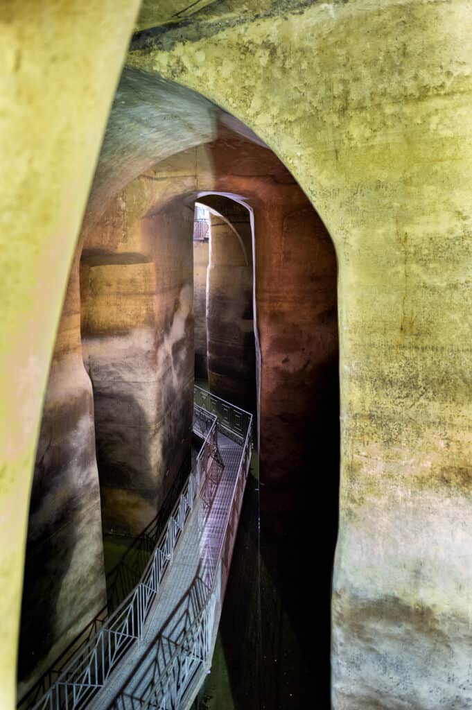 A narrow walkway with railings runs between tall, weathered stone walls, creating an enclosed, cavern-like area with a play of light and shadow.
