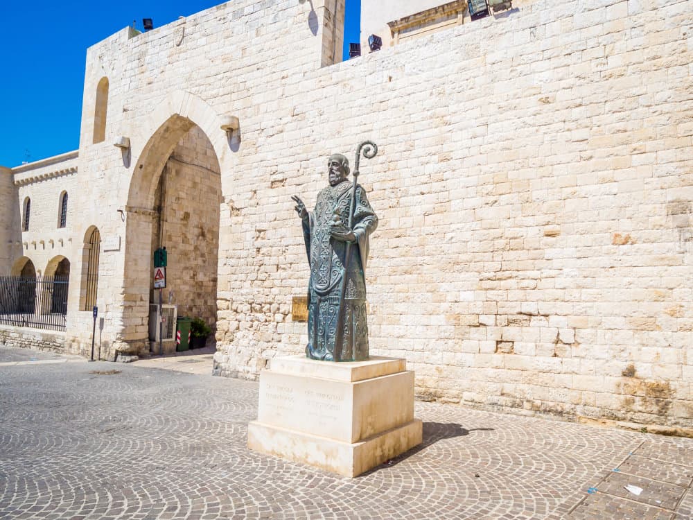 A bronze statue of a robed and bearded man holding a staff stands on a stone pedestal in front of a historic stone building with an arched entryway.