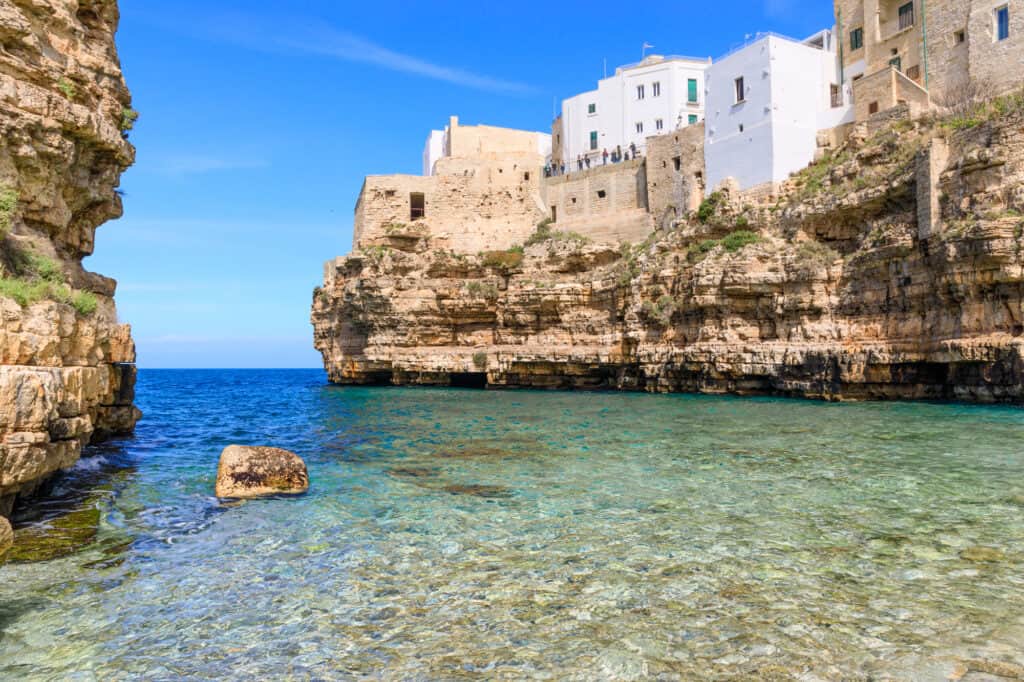 Clear blue water meets rugged cliffs and white stone buildings under a bright blue sky.