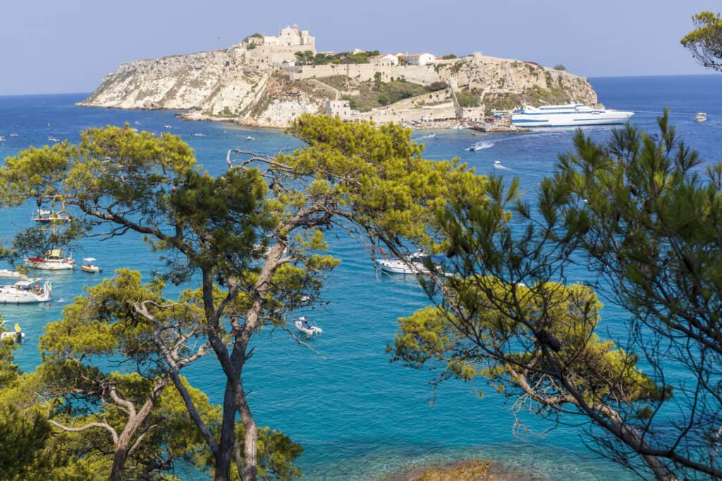 View of a coastal island with a historic fortress on a hill, surrounded by clear blue sea. Several boats are anchored nearby, and pine trees frame the foreground.