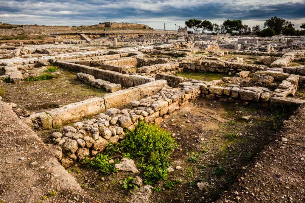 Ancient stone ruins with rectangular structures under a cloudy sky, surrounded by sparse vegetation.