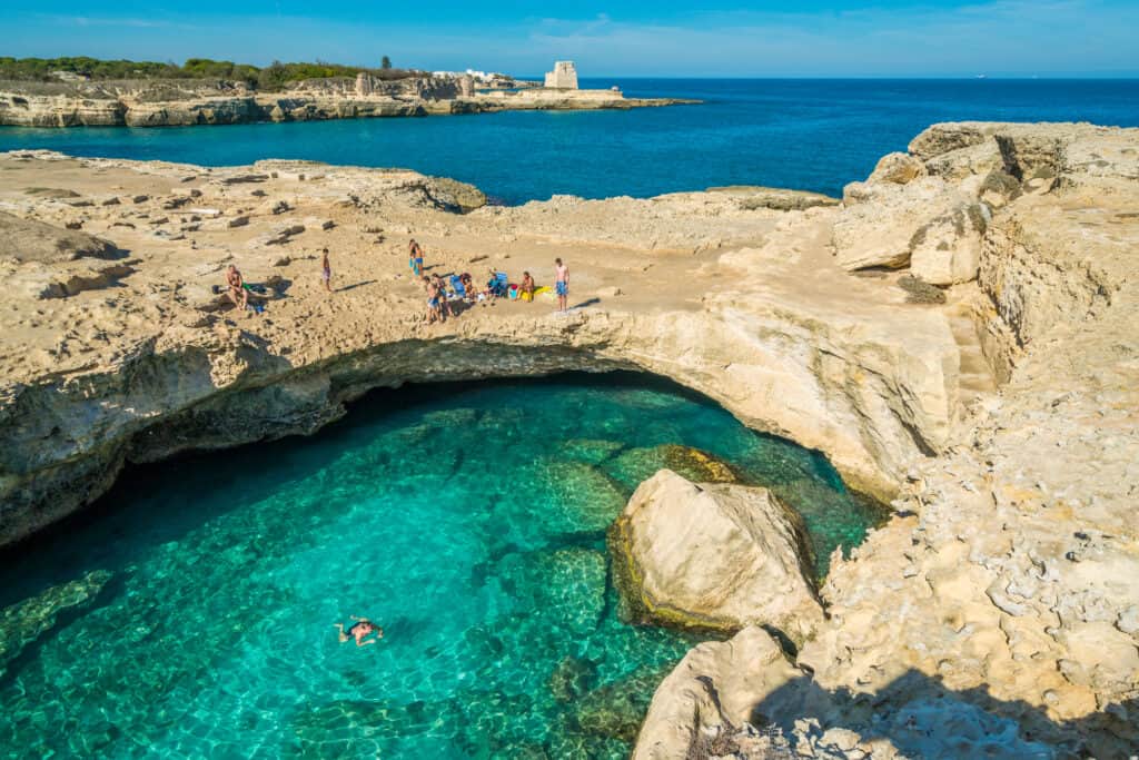 A natural rock formation with a clear blue water pool, surrounded by rocky terrain. People are sitting along the edges under a sunny sky. A tower is visible in the distance.