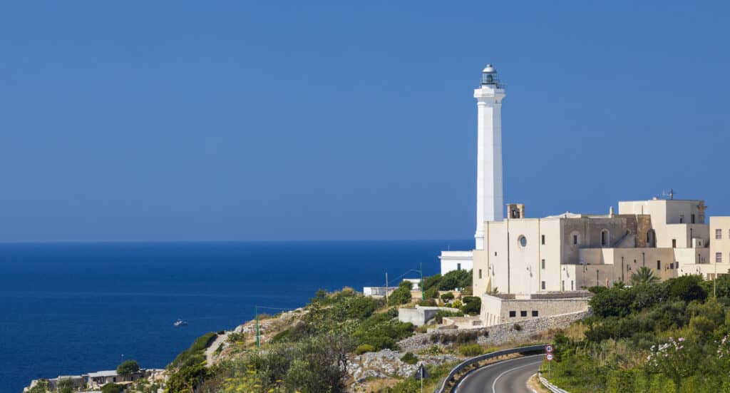 A white lighthouse stands on a coastal cliff with buildings nearby, overlooking a blue sea under a clear sky. A winding road leads towards the lighthouse.