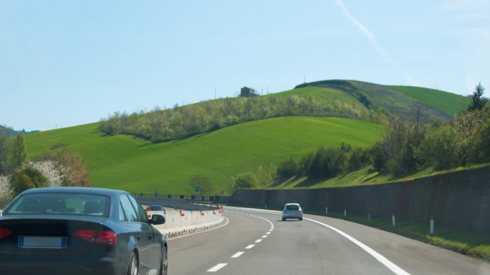 Cars driving on a highway with green hills and a clear blue sky in the background.