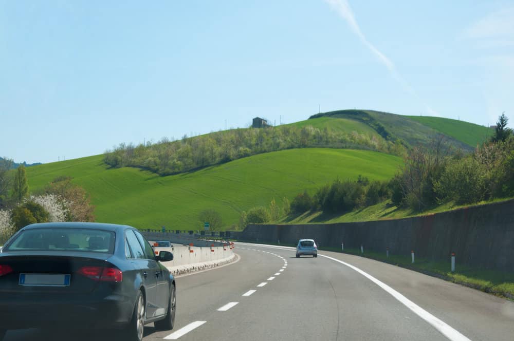 Cars driving on a highway with green hills and a clear blue sky in the background.