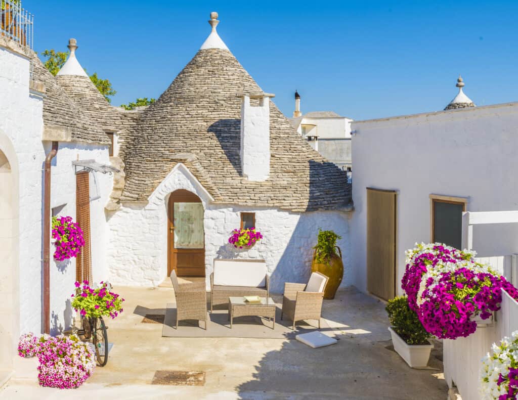 Stone Trullo house with conical roofs in a sunny courtyard, featuring wicker furniture, flowering potted plants, and blue sky in the background.