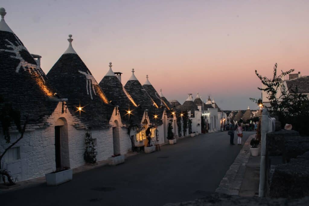 A row of trulli with conical roofs is illuminated at dusk along a quiet street in Alberobello, Puglia, with a few people visible.