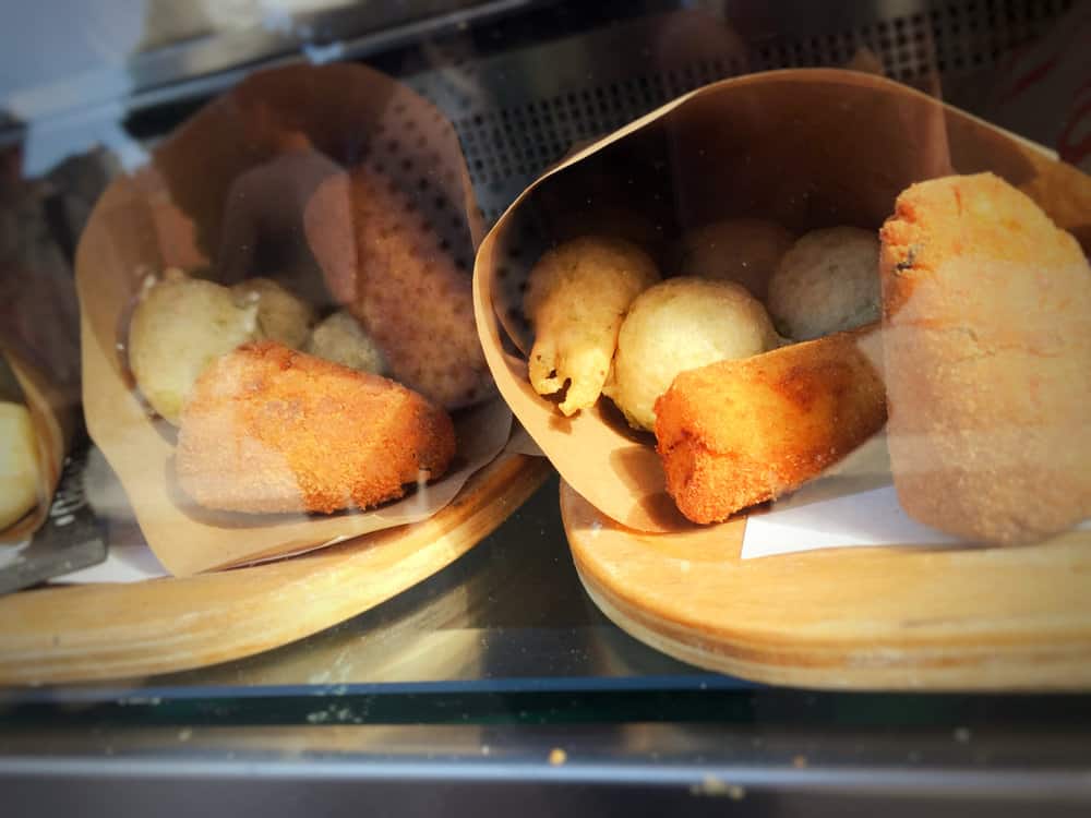 Close-up of two paper containers filled with a delightful assortment of fried foods, reminiscent of the classic Cuoppo from Naples, Italy. The treats are artfully arranged on wooden trays and displayed behind glass.